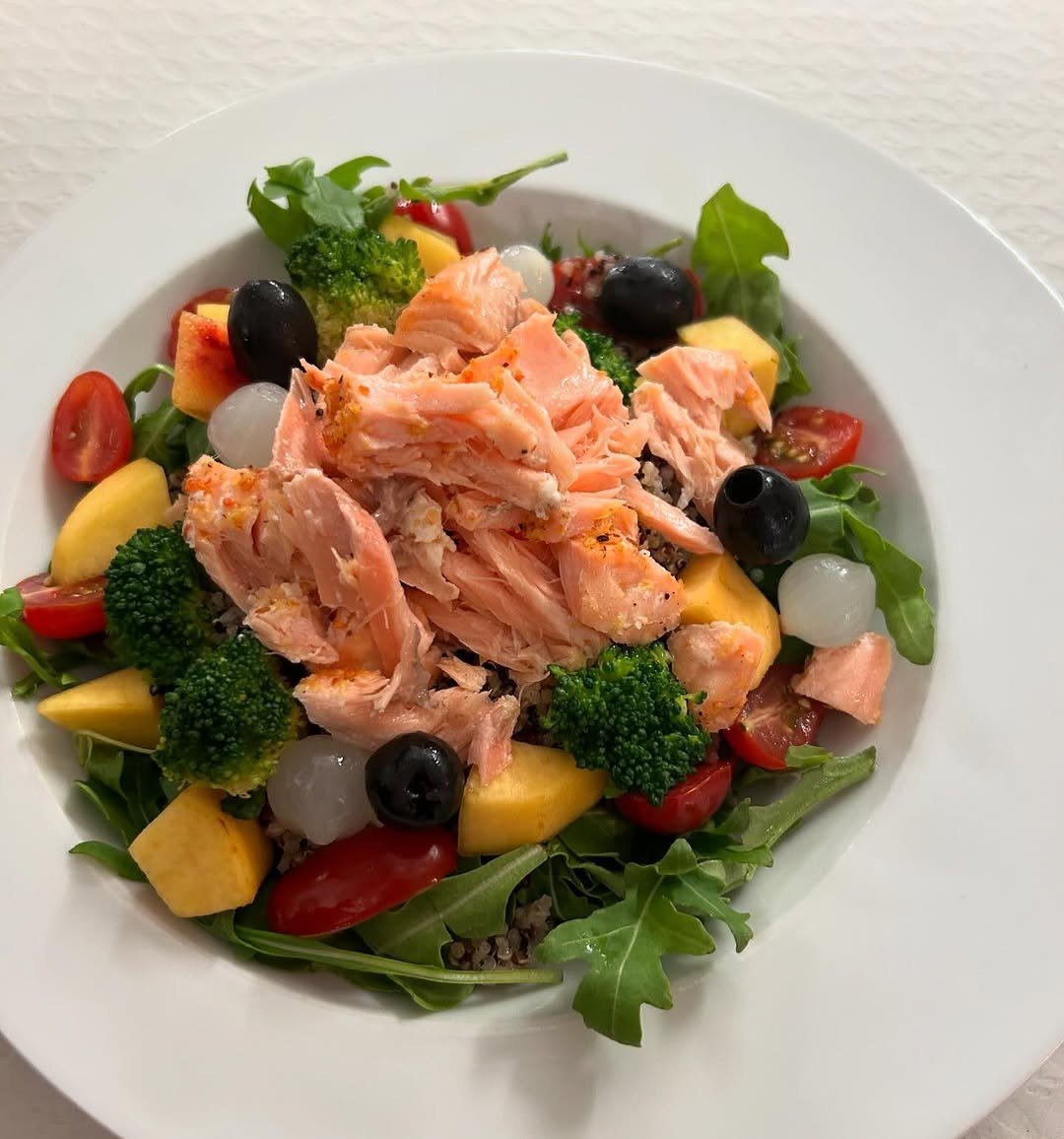 Salada com salmão desfiado, rúcula, brócolos, tomate cereja, azeitonas e cubos de manga num prato branco