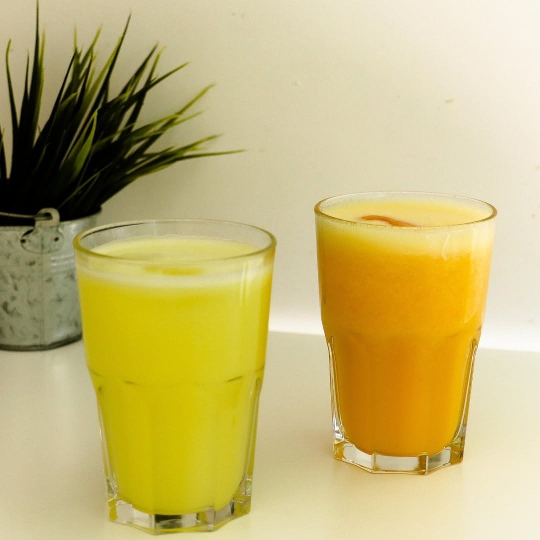 Dois copos de sumo natural, amarelo e laranja, numa mesa clara ao lado de um vaso com uma planta